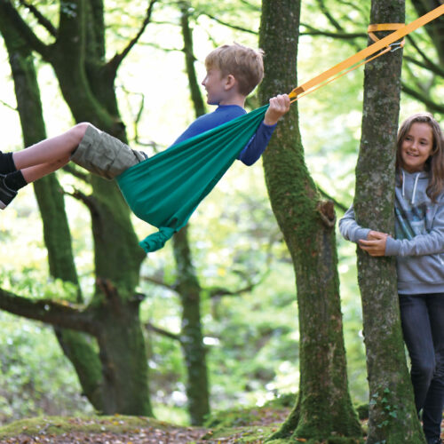 Ein Bub im Schulalter schaukelt in der Taschenschaukel, die im Wald auf einem Baum aufgehängt ist