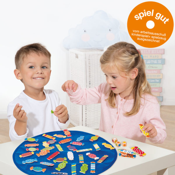 Kinder im Kindergartenalter spielen an einem Tisch mit dem Würfelspiel Candy. Zusätzlich abgebildet ist das Spiel Gut Logo
