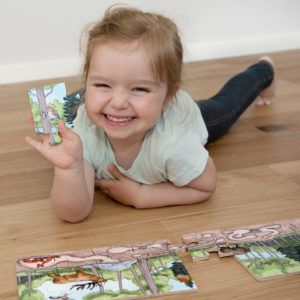 Mädchen im Kindergartenalter liegt am Boden und setzt das Puzzle Entdecke den Wald zusammen