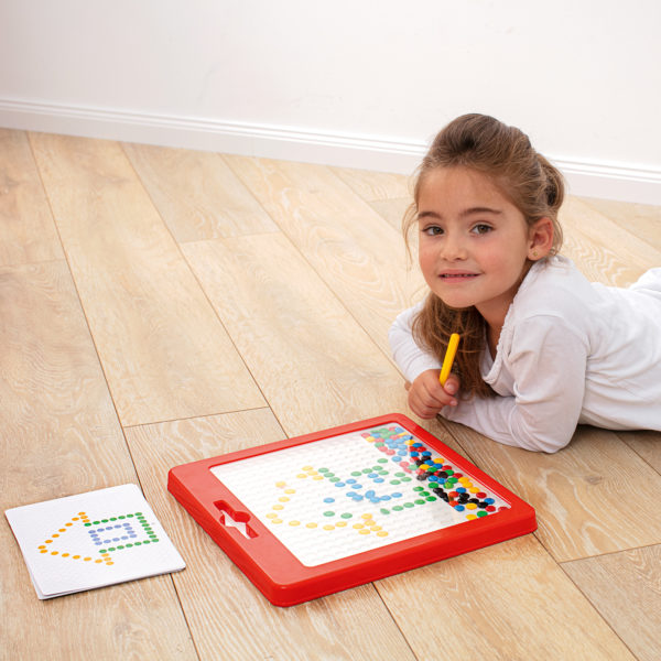 Mädchen im Kindergartenalter liegt am Boden und zeichnet mit dem Magnetstift des Magnetspiels Kunterbunt eine Vorlagenkarte nach
