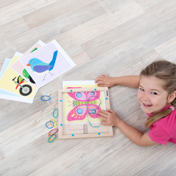 Mädchen im Kindergartenalter liegt am Boden und zeichnet mit den Gummibändern des Flexipainters auf dem Holzrahmen die Schmetterlingsvorlage nach. Außerdem zu sehen: einige andere Vorlagenblätter. unterbunt eine Vorlagenkarte nach