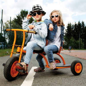 Zwei Kinder im Kindergartenalter sitzen in cooler Pose mit Sonnenbrille auf dem Dreirad "Taxi" von beleduc in orange