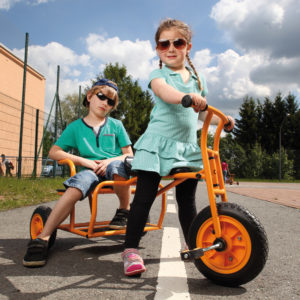 Zwei Kinder im Kindergartenalter sitzen in cooler Pose mit Sonnenbrille auf dem Dreirad "Doppeltaxi" von beleduc in orange