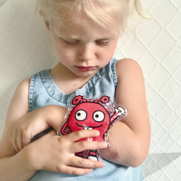 Mädchen im Kindergartenalter hält sich ein rotes Helfermonster als Coolpack auf den verletzten Arm.