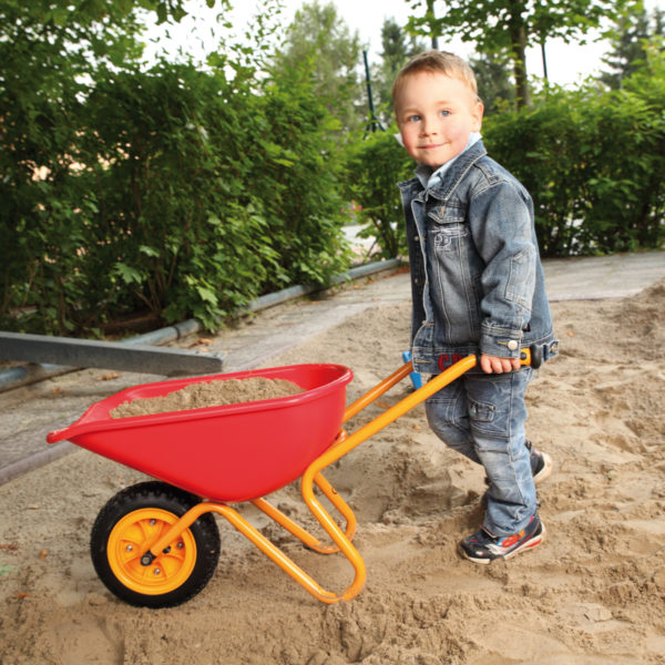 Bub im Kindergartenalter schiebt die leichte und robuste Scheibtruhe mit Sand beladen durch die Sandkiste