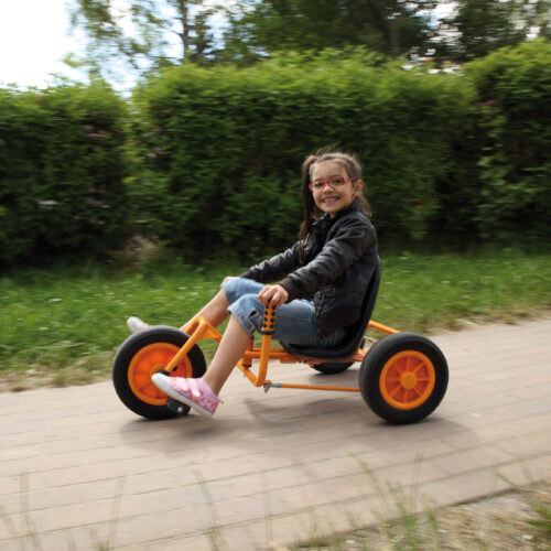 Kind im Volksschulalter saust mit dem Dreirad Rider, das über einen niedrigen Sitz und eine beidseitige Lenkvorrichtung in Höhe des Sitzes verfügt, durch den Garten.