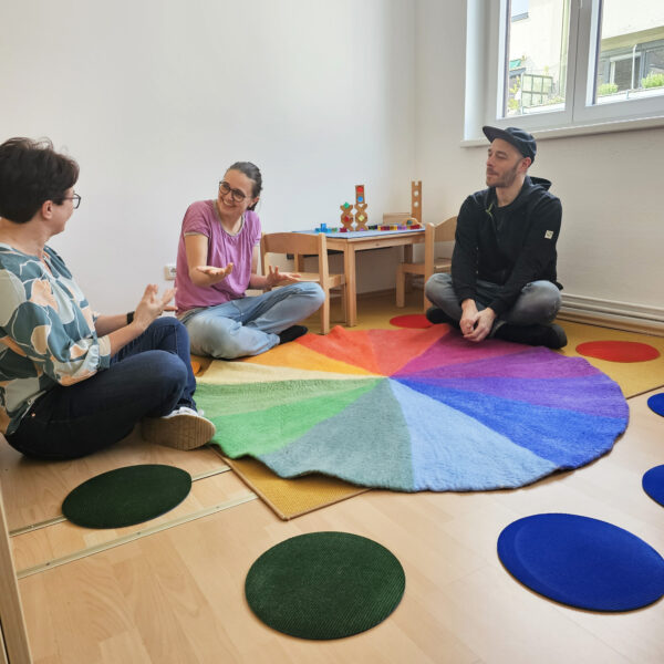 3 Erwachsene sitzen um den Farbenkreis als Mitte auf den runden Teppichfliesen
